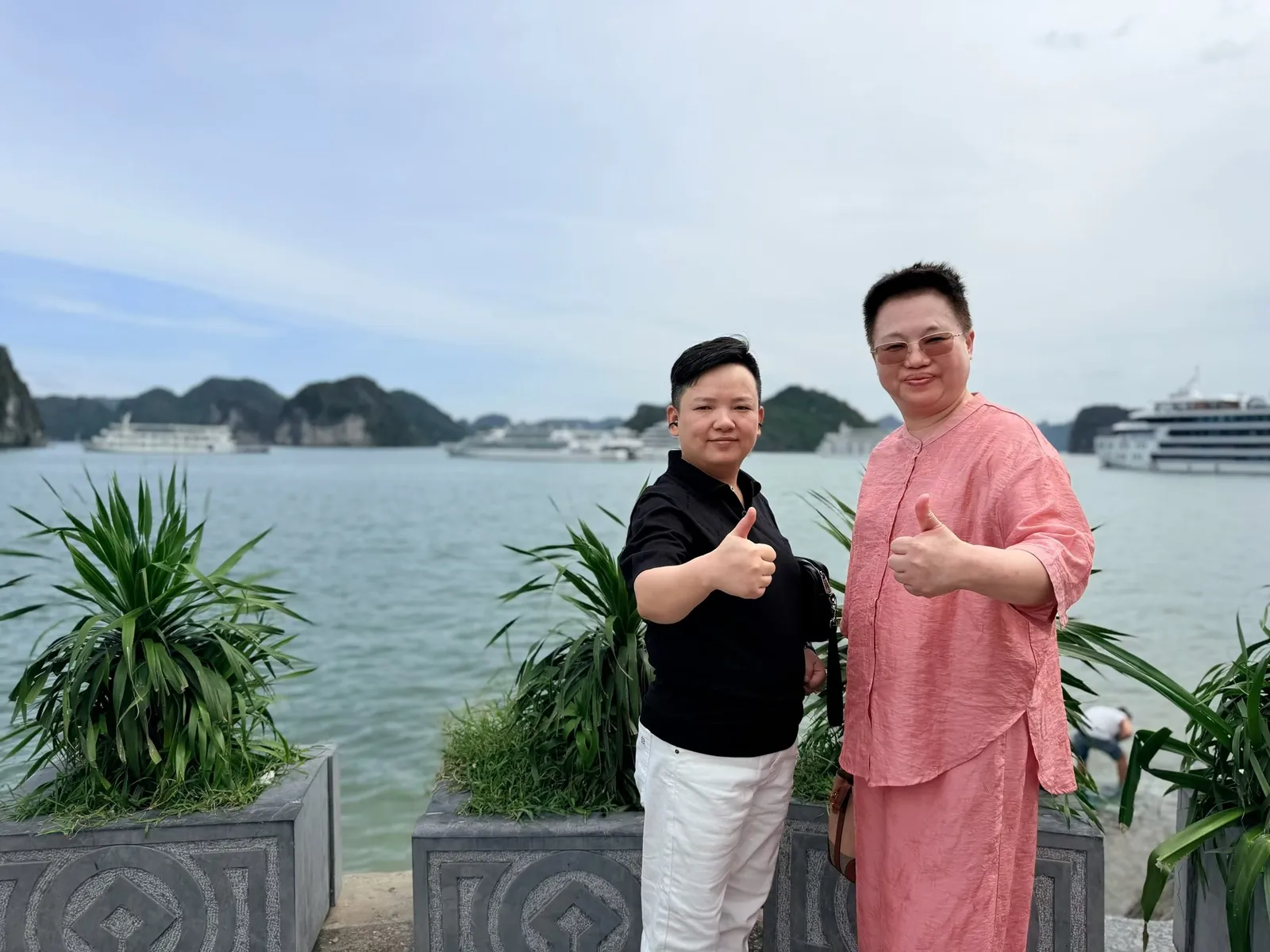 两人在下龙湾岸边竖起大拇指,身后是海面、游船和远处的群山